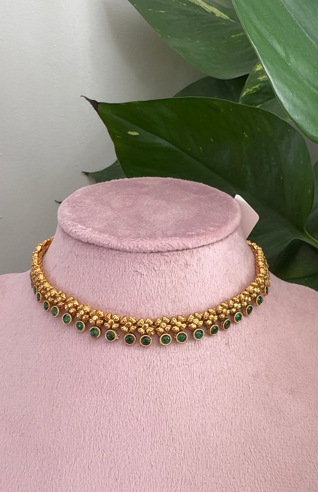 Gold necklace with green stones on a pink mannequin against a green leafy background