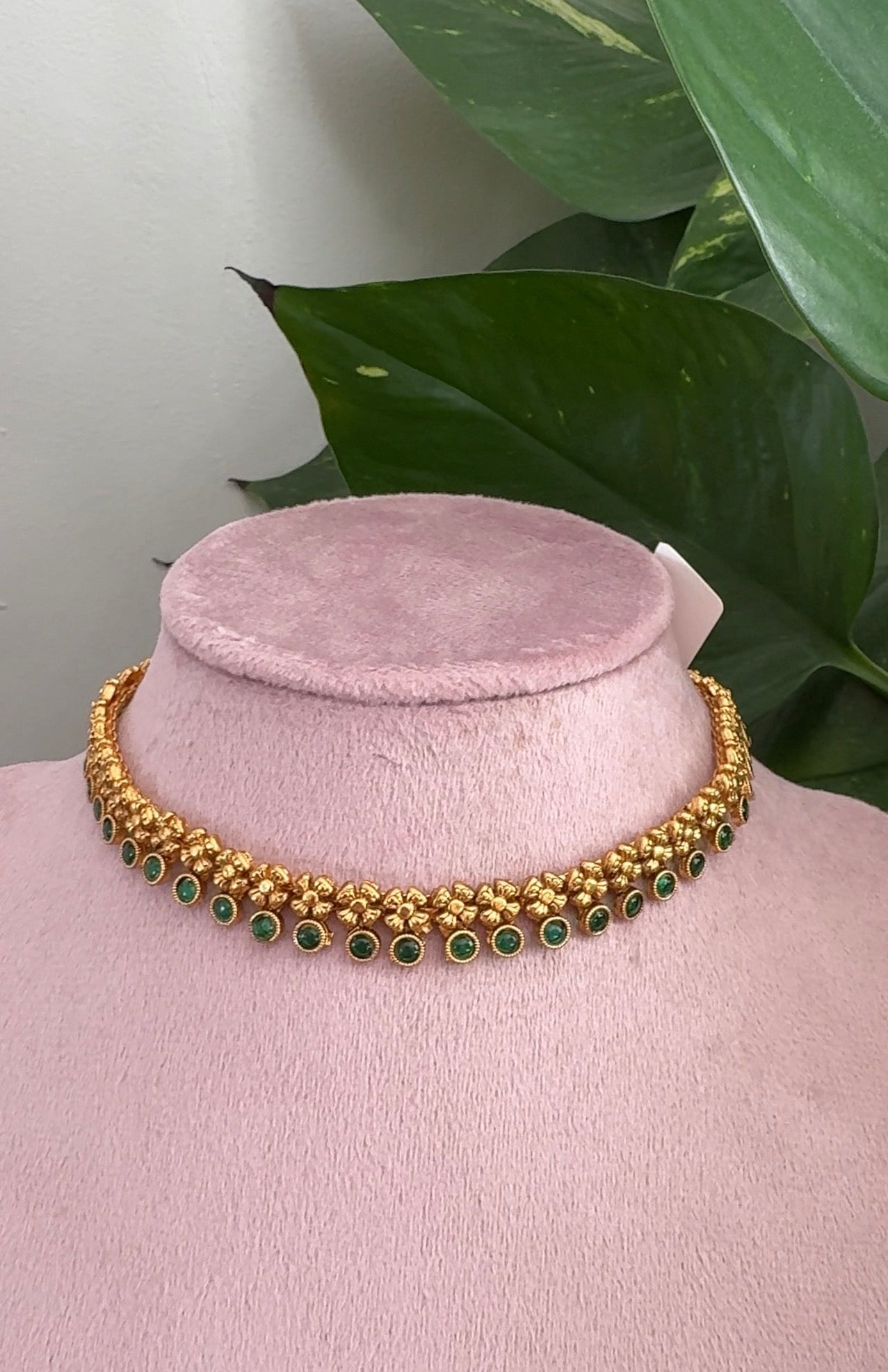 Gold necklace with green stones on a pink mannequin against a green leafy background